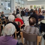 Skupina stanovalcev sedi v dvorani, ploska in spremlja nastop glasbenikov s harmoniko, klarinetom, kitaro in kontrabasom.