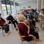 Skupina stanovalcev sedi na stolih in invalidskih vozičkih ter spremlja glasbeni nastop.