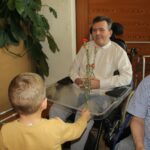 Deček v rumeni majici izroča rdeč nagelj moškemu na invalidskem vozičku, ob njiju stojita še en deček in moški.
