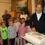 Moški z nožem reže veliko pravokotno torto, ob njem pa stoji skupina otrok.