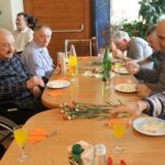 Skupina stanovalcev sedi za mizami s krožniki torte, kozarci pijače in rdečimi nageljni.