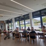 Skupina oseb sedi za mizami in posluša predstavitev ob projekcijskem platnu.