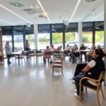 Skupina oseb sedi v prostoru med predstavitvijo, dve osebi stojita ob projekcijskem platnu.