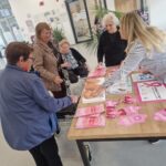 Več oseb stoji ob mizi z rožnatimi informativnimi gradivi in modeloma dojk.