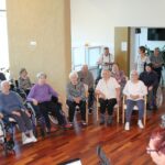 Skupina ljudi sedi v polkrogu in posluša moškega, ki igra harmoniko.