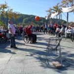 Moški na vozičku meče košarkarsko žogo proti košu, medtem ko ga opazuje skupina ljudi.