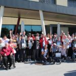 Večja skupina ljudi, nekateri na invalidskih vozičkih, pozira pred stavbo in drži priznanja.