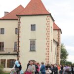 Skupina ljudi, nekateri na invalidskih vozičkih, se v koloni premika mimo grajske stavbe.