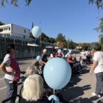 Skupina ljudi z modrimi baloni spremlja osebe na invalidskih vozičkih po cesti.