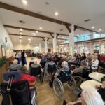 Večja skupina ljudi, nekateri na invalidskih vozičkih, sedi v dvorani in spremlja nastop harmonikarjev.