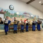 Skupina harmonikarjev v enakih oblačilih stoji v vrsti in igra harmonike.
