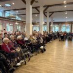 Večja skupina ljudi, nekateri na invalidskih vozičkih, sedi v dvorani in spremlja harmonikarja.