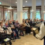 Skupina sedečih ljudi poje in ploska med nastopom harmonikarja.