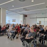 Obiskovalci sedijo v dvorani in spremljajo dogajanje pred njimi.