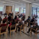 Predavateljica z mikrofonom govori sedeči skupini v dvorani, za njo sta projekcijska zaslona.