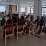 Predavateljica z mikrofonom nagovarja sedeče udeležence pred projekcijskima zaslonoma.